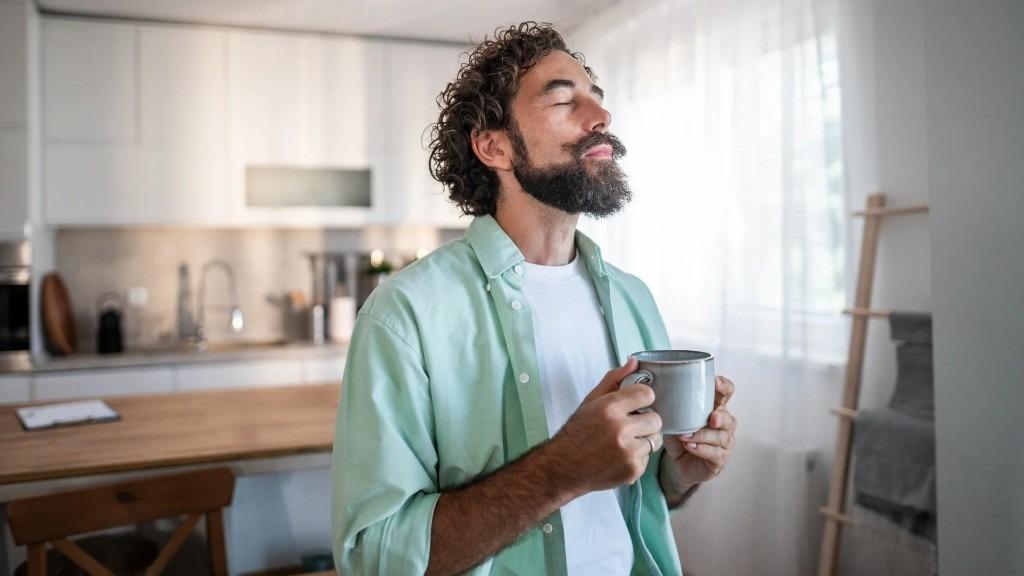 A man in a white tee and open mint shirt holding a mug in a sunlit kitchen.